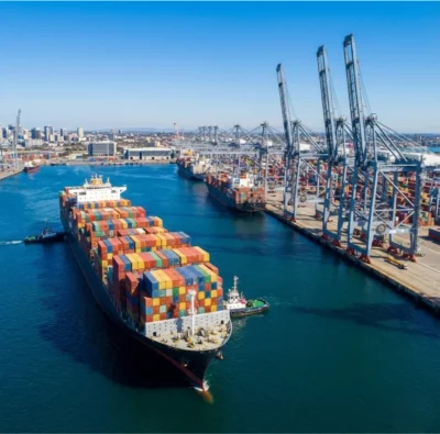global-shipping-port-logistics High-angle view of a large cargo ship and tugboat arriving at a busy shipping terminal with gantry cranes and a city skyline.